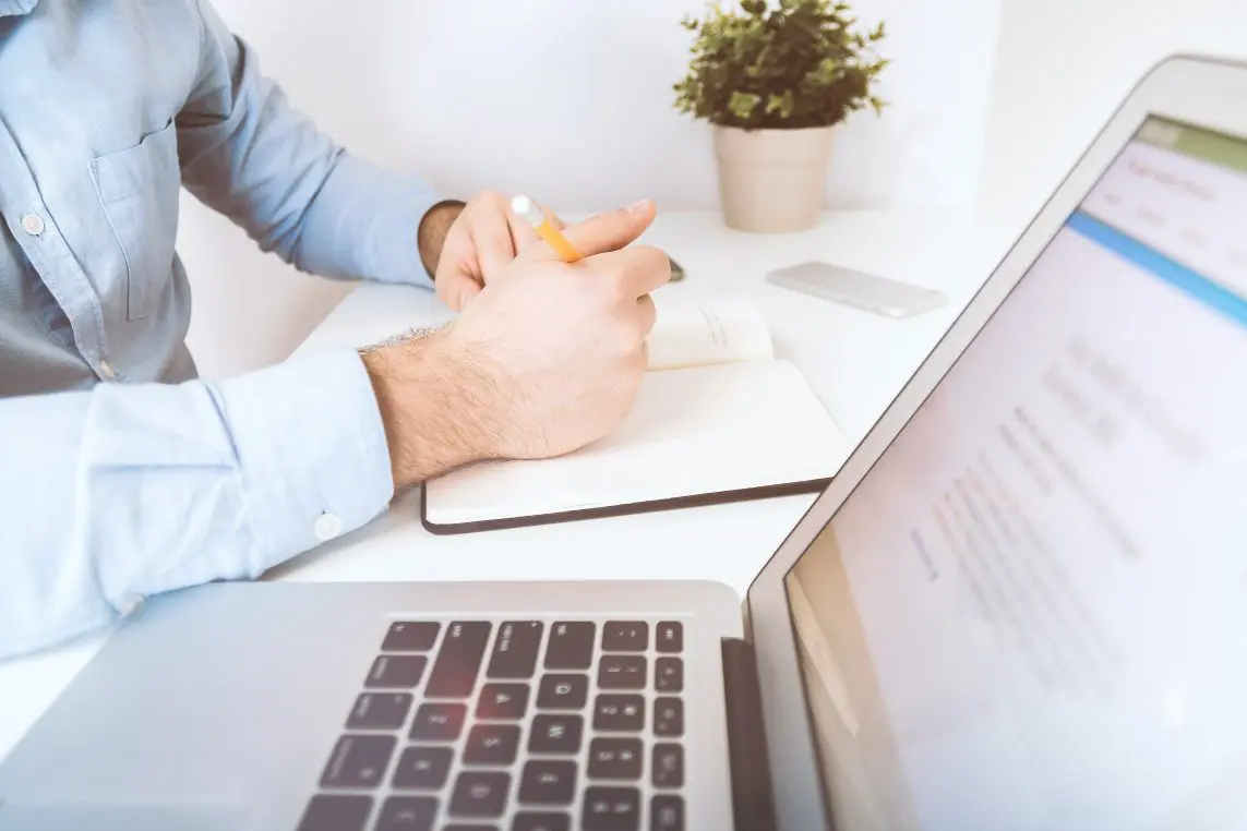 Person's hands write with a pencil in an open notebook while a laptop shows a blurred webpage (text unreadable) and a small potted plant sits on a white desk.