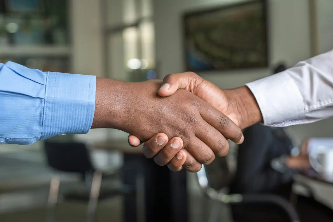 Two hands clasp in a handshake, one from a light-blue shirt, the other from a white dress shirt, set in a blurred office with chairs and framed artwork.