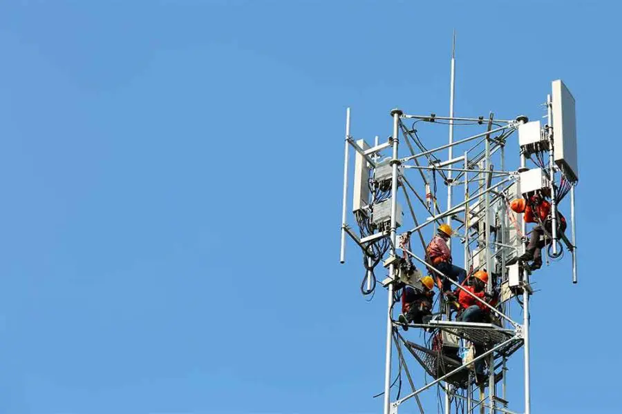 Telecommunications lattice tower with several technicians wearing orange vests, helmets, and harnesses, climbing and installing antennas and thick cables high above the ground against a clear blue sky.