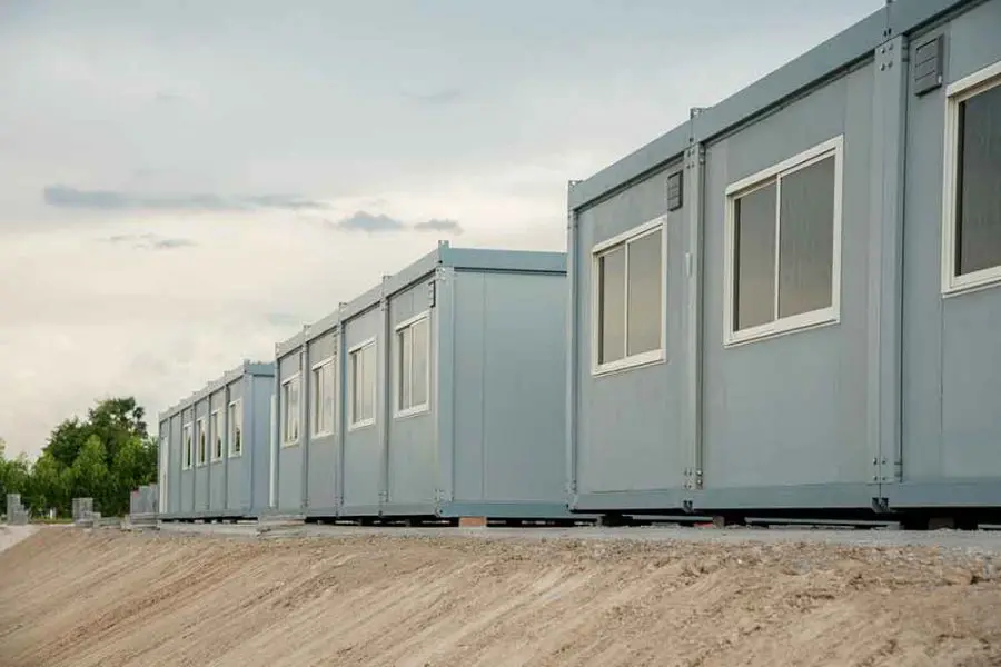 Gray modular container units line up on a raised gravel embankment, their windows facing outward, set against an overcast sky with distant trees.