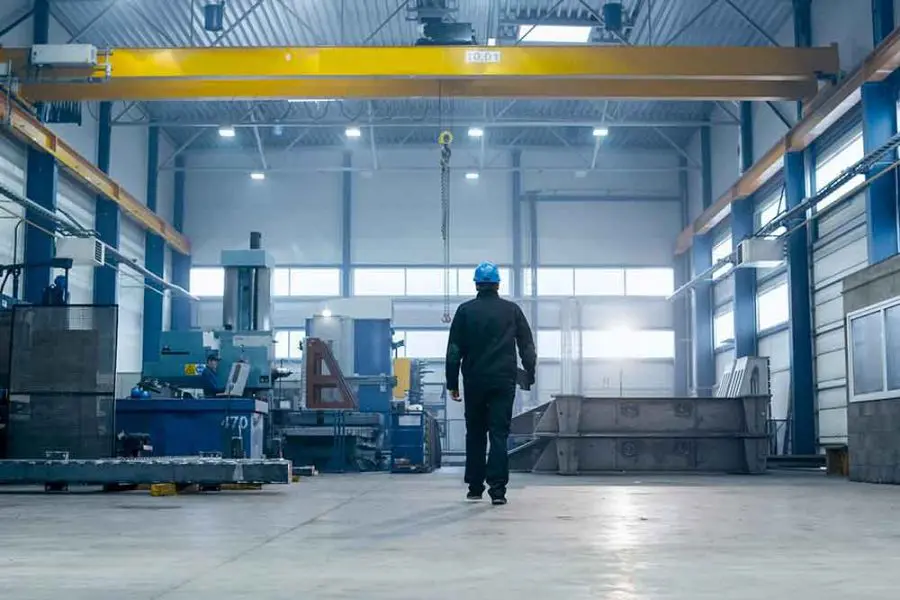 Worker in a blue hard hat walks toward a suspended yellow overhead crane labeled 