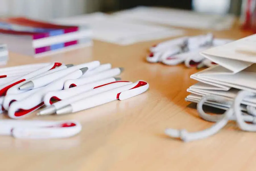 White plastic pens with red accents lie scattered across a wooden table, near a stack of folded paper bags and coiled lanyards, with blurred notepads and materials in the background.