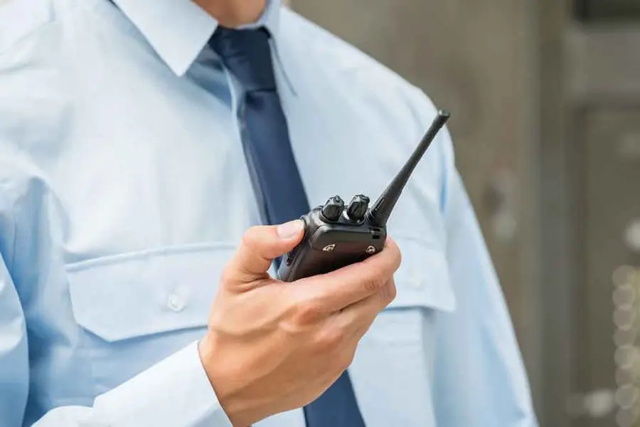 A person in a light-blue uniform shirt and navy tie holds a black walkie-talkie at chest level, manipulating its knobs; blurred gray metal panels in the background suggest an indoor facility.