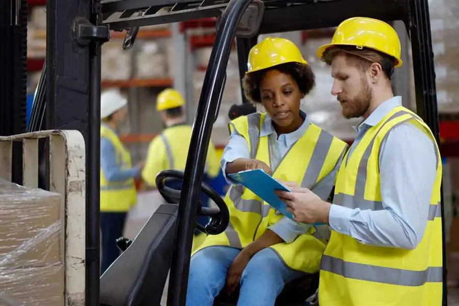 Forklift and workers: a seated woman points to a clipboard the standing man holds; both wear hard hats and high-visibility vests in a warehouse with stacked pallets and colleagues.