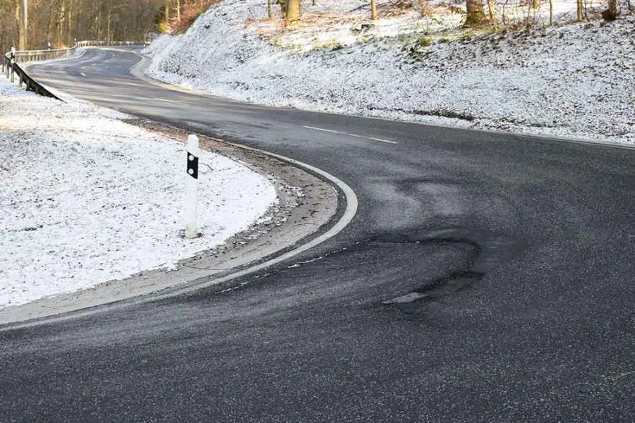 A winding asphalt road curves left, displaying dark skid marks and surface patches; a white reflector post marks the inner bend, surrounded by snow-covered shoulders and a sparsely wooded roadside.