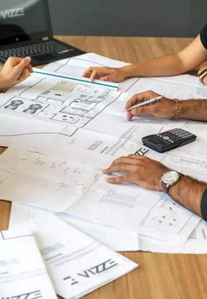 Architectural blueprints spread on a wooden table; hands measure and annotate with a ruler and pen while a calculator and laptop sit nearby. Text visible: 