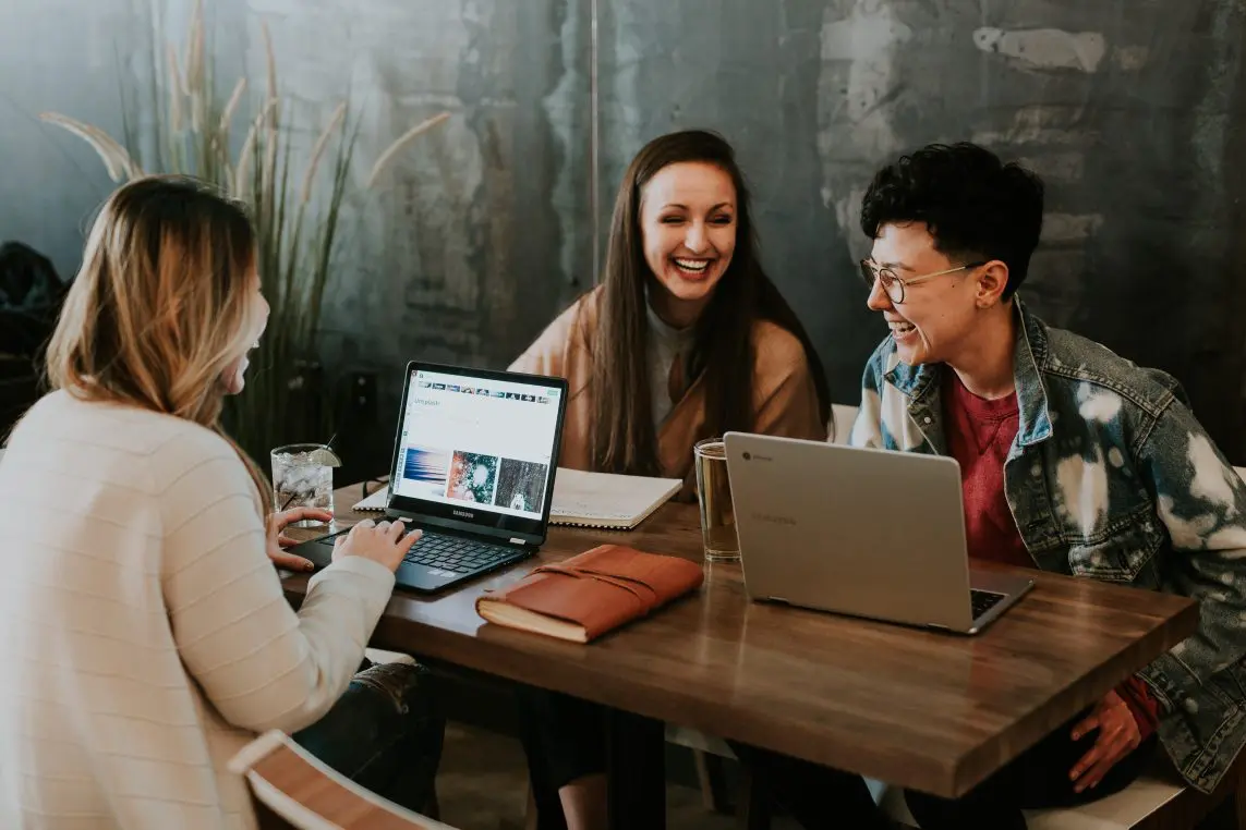 Three young adults sit around a wooden table, sharing laptops and notebooks, laughing together while working in a cozy café-like space with plants and a dark textured wall.