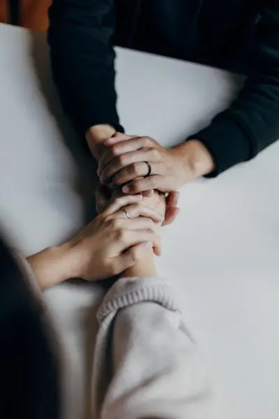 Two pairs of hands clasping—one hand gently covering the other—resting atop a white table; one person in a dark long sleeve, the other in a light sweater; both wear simple rings.