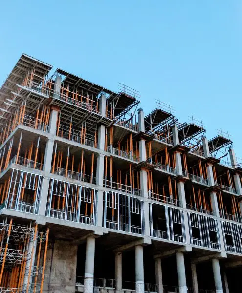 Multi-story concrete building under construction, exposed columns and steel framing with orange vertical formwork posts and scaffolding rising across multiple floors against a clear blue sky.