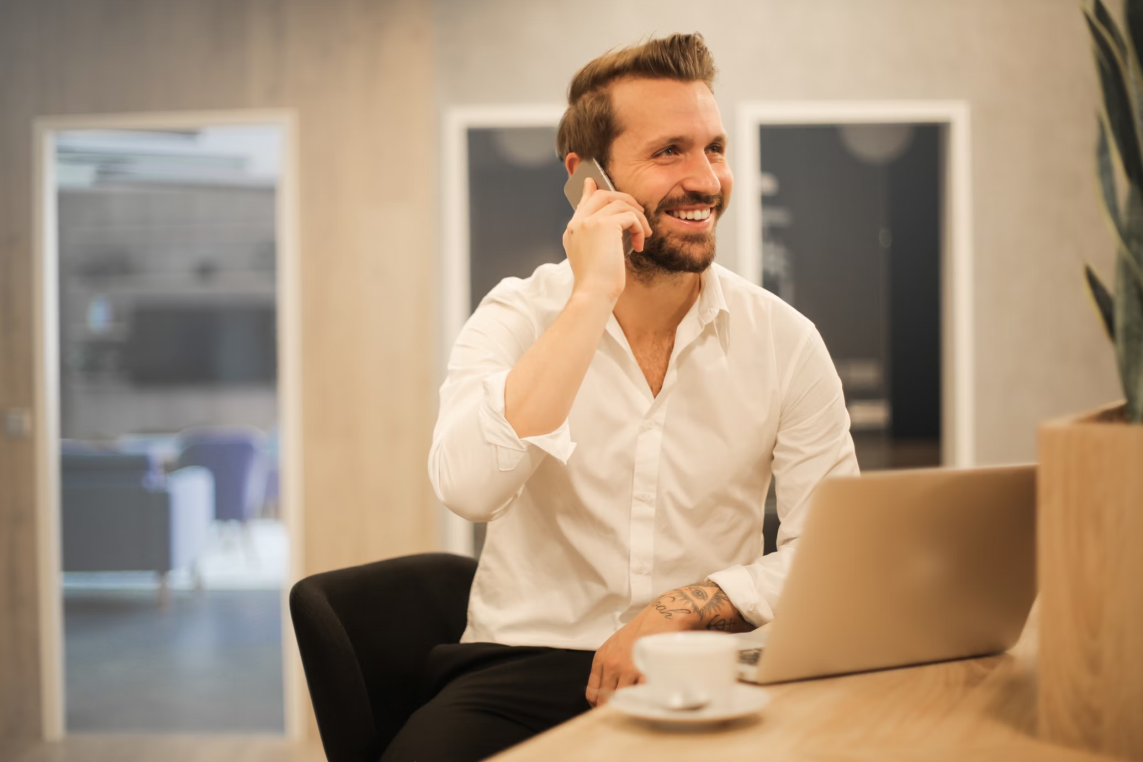 Man in a white shirt talks on a smartphone while seated at a desk with an open laptop and coffee cup in a modern office lounge.