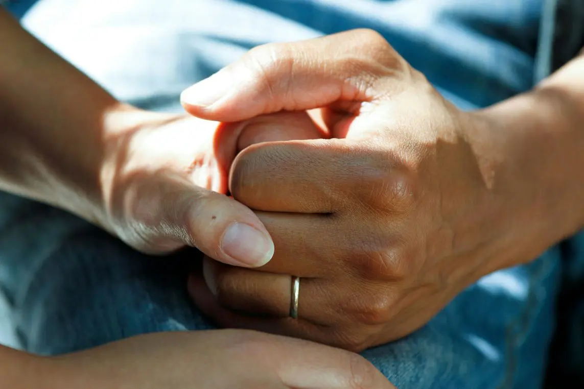 Two adult hands — one wearing a simple gold band — clasp each other gently, offering comfort while resting on a denim-clad lap in warm, dappled sunlight.