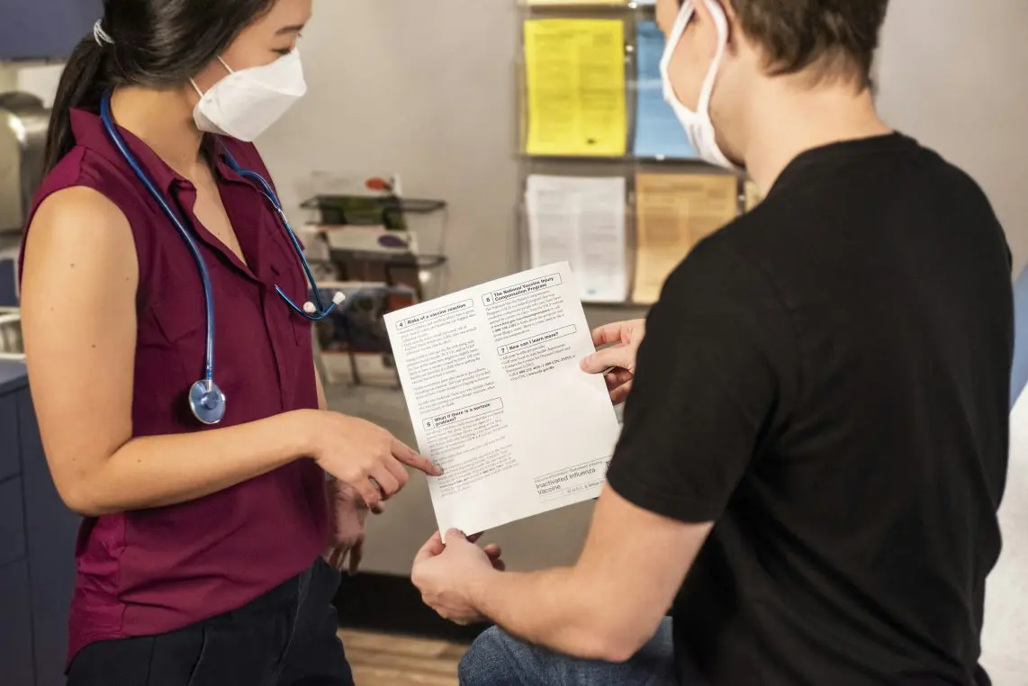 Medical professional wearing a mask and stethoscope points to a printed information sheet held by a masked patient; they consult in a clinic room with a bulletin board and supplies in the background. Printed text on the sheet is not legible in the photo.