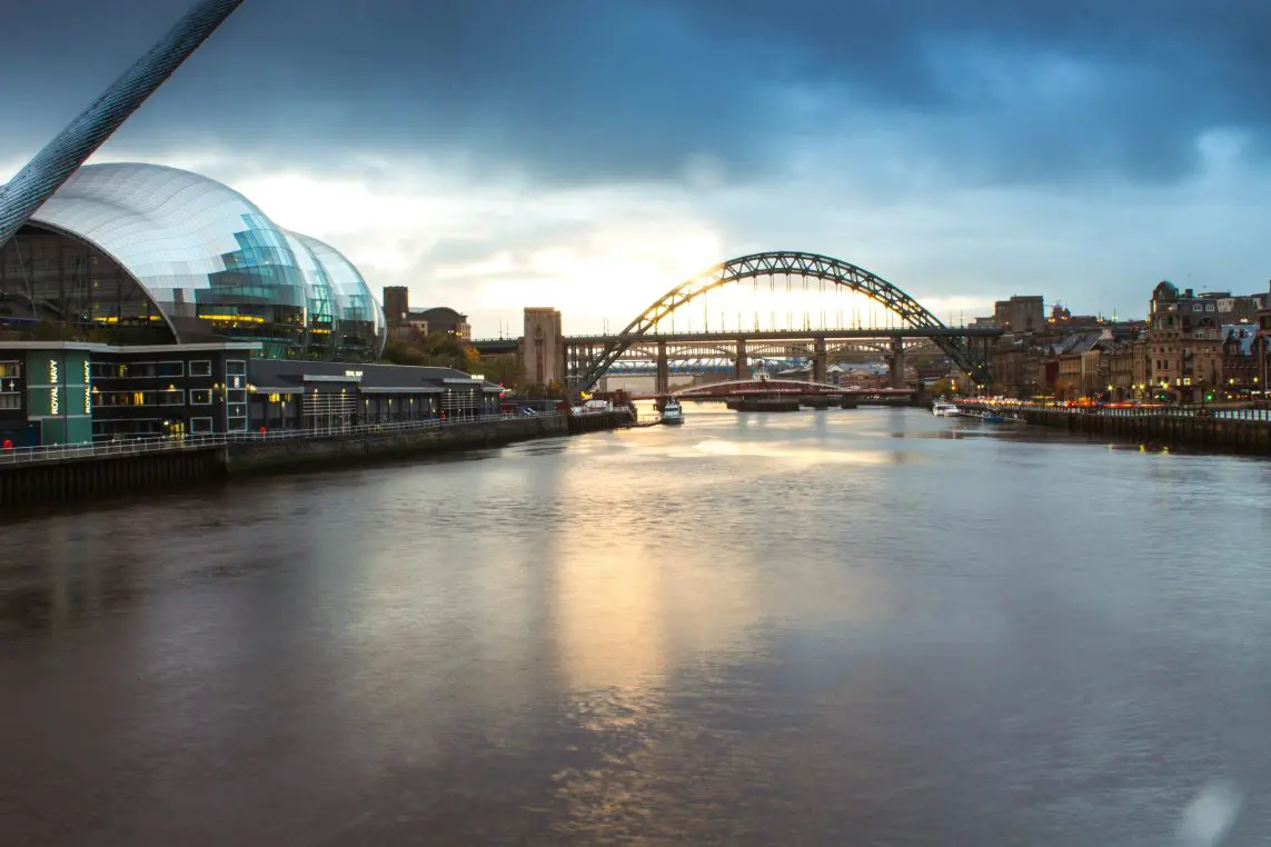 Tyne River flows gently; curved glass performance venue hugs the left bank while multiple arched bridges span the water; twilight sky and city lights reflect across the calm river.
