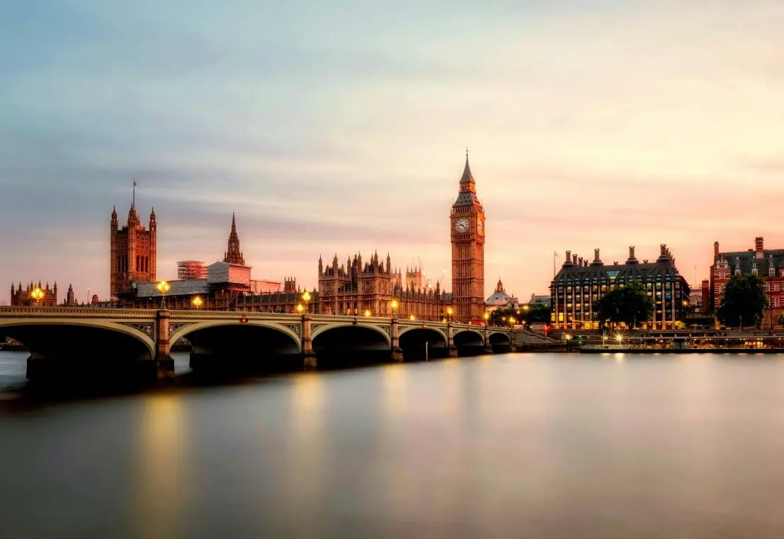 Elizabeth Tower (Big Ben) and the Palace of Westminster rise, illuminated, beside Westminster Bridge, their lights reflecting on the calm River Thames at sunset under a pastel sky.