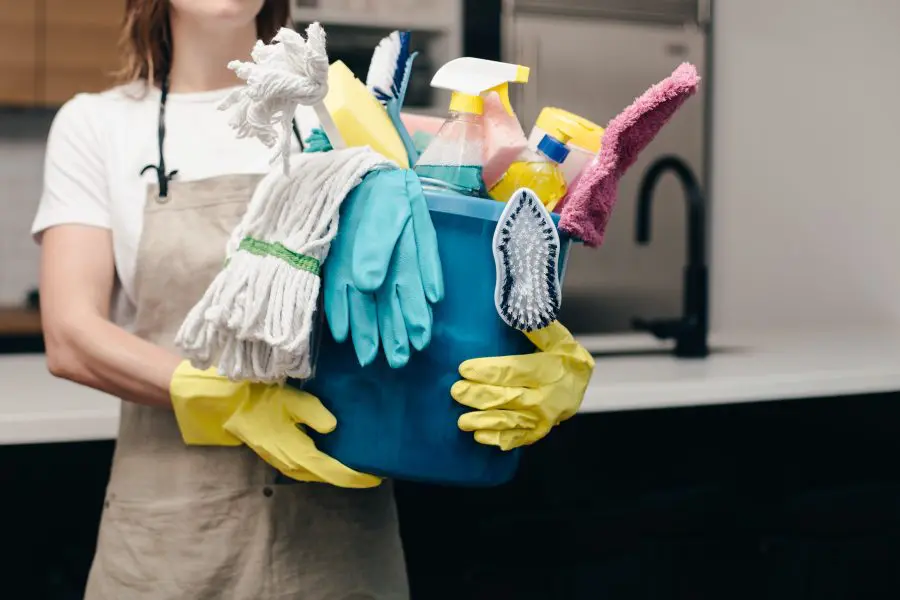 Blue plastic bucket filled with cleaning supplies (mop head, spray bottles, sponge, scrub brush, rubber gloves) is held by a person wearing yellow rubber gloves in a modern kitchen.