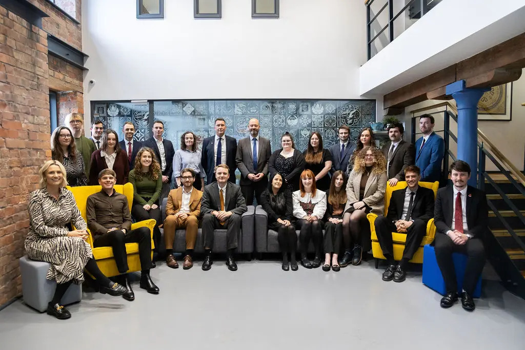 About two dozen professionals pose for a formal portrait, seated and standing in rows in a modern office lounge with exposed brick, patterned glass panel, mezzanine and yellow armchairs.