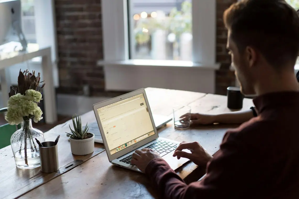 Laptop on a wooden table being typed on by a seated man; sunlight through a window illuminates a brick-walled workspace with a vase of flowers, small succulent, cup and glass.