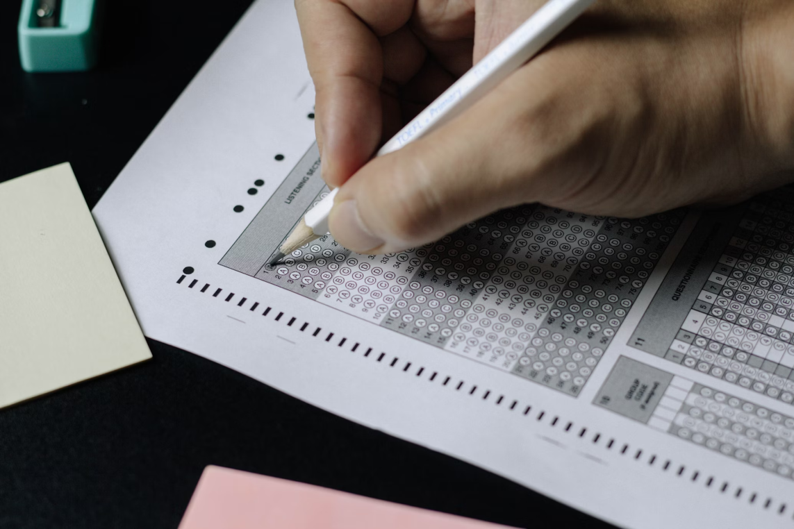 A hand fills bubbles on a multiple-choice answer sheet with a pencil, placed on a dark desk beside sticky notes and a green pencil sharpener.