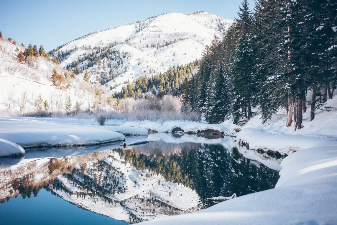 Snow-covered mountain reflected in a calm river, framed by snow-laden banks and evergreen trees; sunlight highlights ridges and scattered pines beneath a clear blue sky.