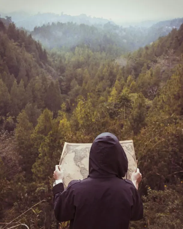 Person in a dark hooded jacket holding an unfolded map, standing at a forested overlook, misty tree-covered valley stretching into the distance.