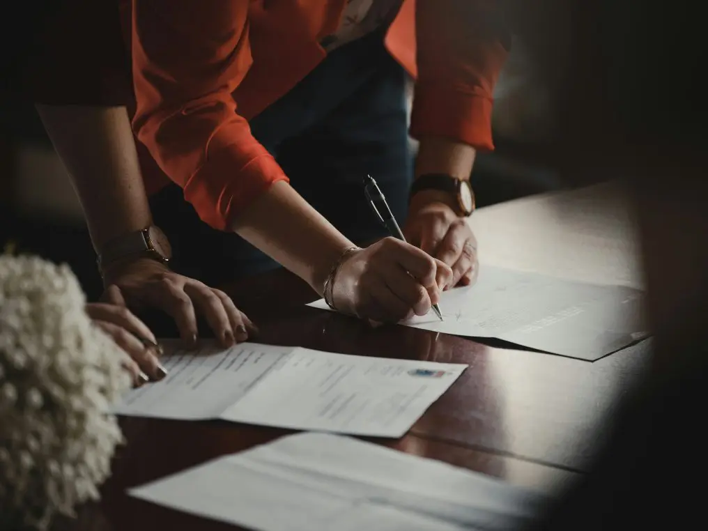 Hands signing documents with a pen, another hand steadying papers on a wooden table; person leans over papers in a dimly lit office or meeting setting.