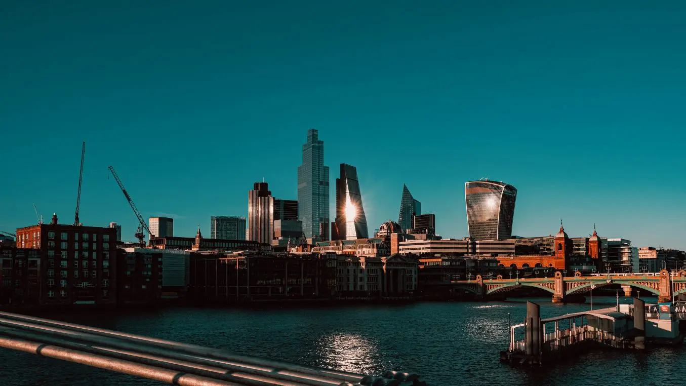 Modern skyscrapers reflect bright sunlight along a riverfront, framed by a bridge, docks, cranes, and a clear teal sky.