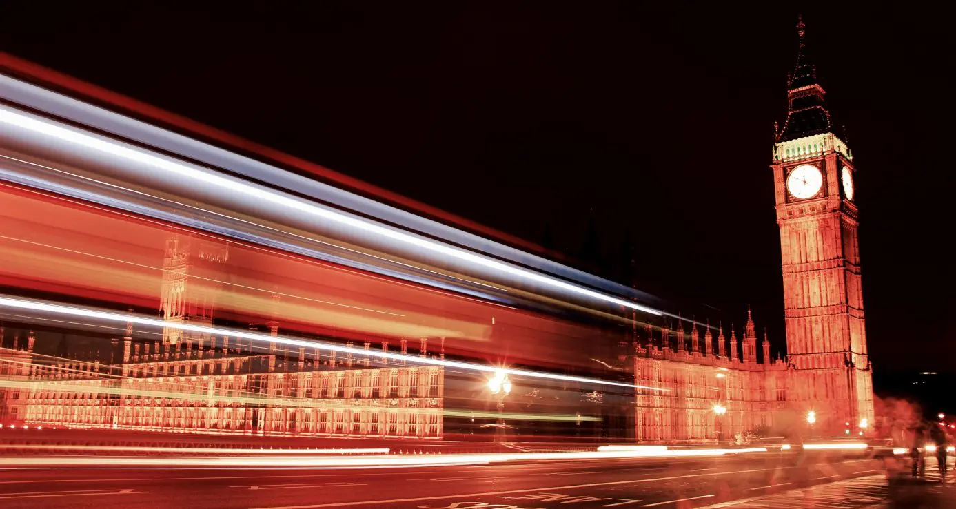 Clock tower and adjacent Gothic parliament building stand illuminated while red bus light trails streak horizontally across a nighttime London bridge roadway.