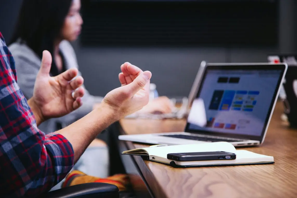 A person's hands gesture expressively over a conference table; a laptop displays blurred charts, a notebook and smartphone rest nearby, colleagues blurred in the background.