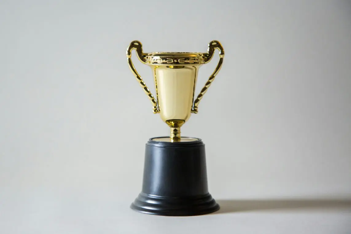 A small gold trophy cup with two ornate handles stands on a black pedestal, centered on a neutral light-gray background and casting a faint shadow to the right.