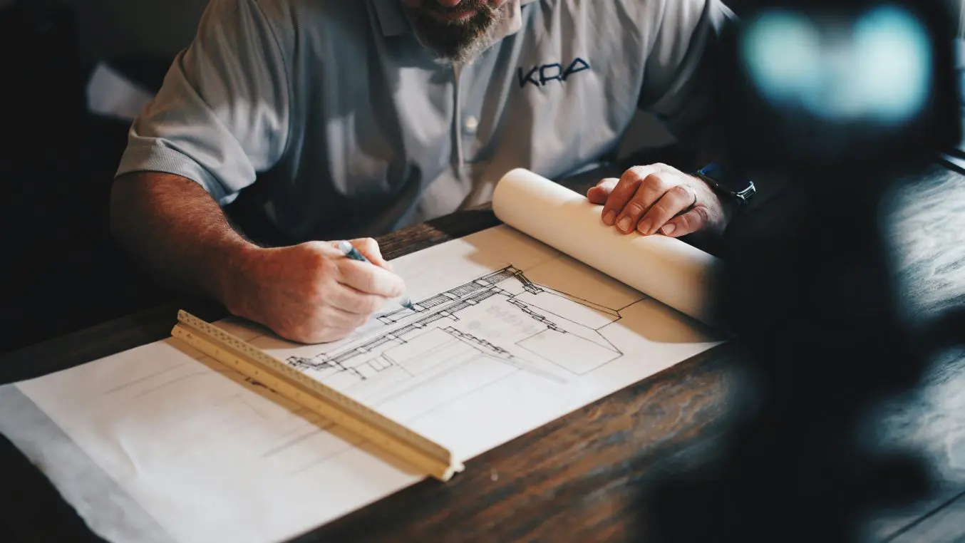Architectural drawing being sketched and annotated by a man with pen on a wooden table, rolled plans and triangular scale rule beside him in a dim workspace. Shirt logo: KRA.