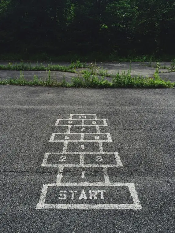 Painted hopscotch grid on cracked asphalt leading toward overgrown weeds and trees, numbered ascending toward the far edge. Text: START; 1, 2, 3, 4, 5, 6, 7, 8, 9, 10.