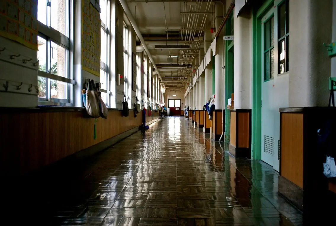 A long school hallway stretches into the distance, sunlight through tall windows casting reflections on the shiny floor; classroom doors line the right, bags on hooks hang along the left.