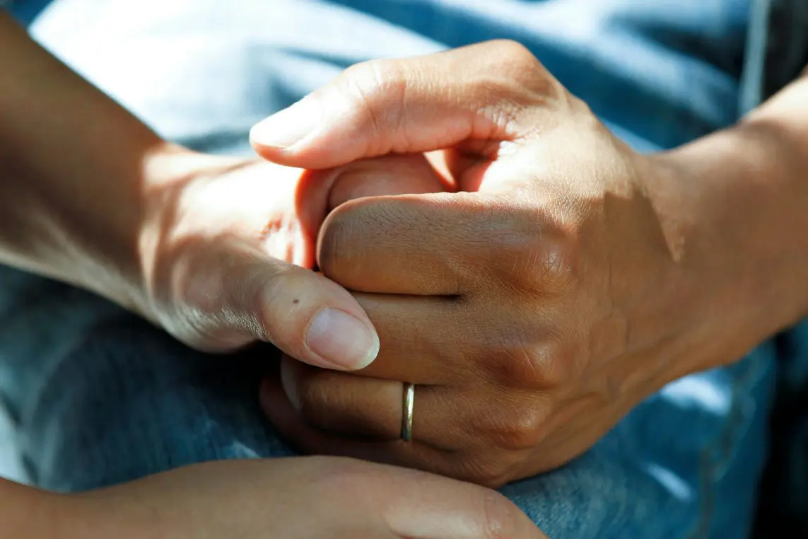 Two adult hands — one wearing a simple gold band — clasp each other gently, offering comfort while resting on a denim-clad lap in warm, dappled sunlight.