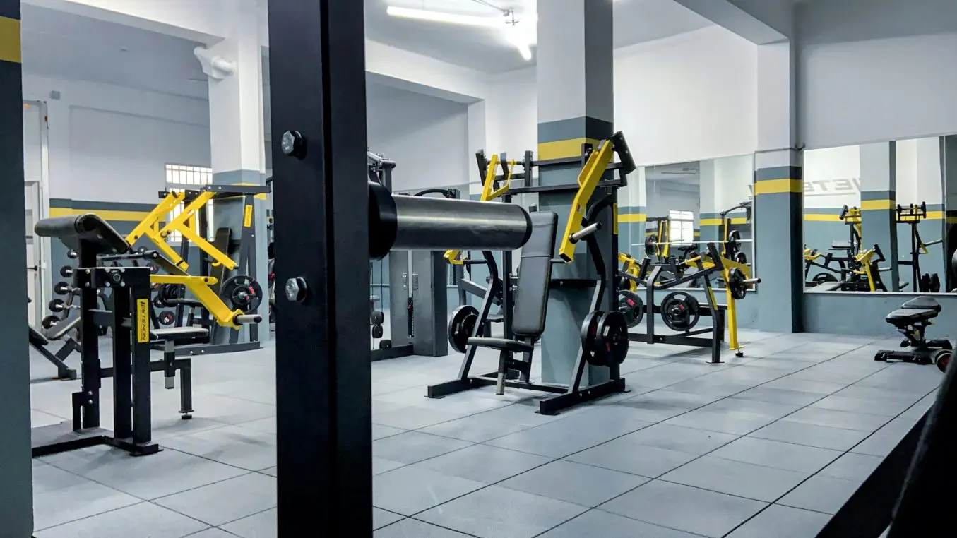 Weight machines stand idle across a bright, modern gym floor with gray tiles, mirrored walls, and yellow-accented equipment reflecting throughout the spacious workout area.