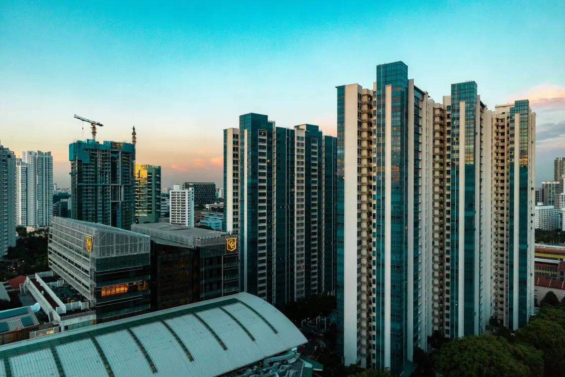 Cluster of tall residential high-rises rise against a dusky sky, flanked by a construction crane and mid-rise buildings, overlooking a curved‑roof complex within a dense urban skyline.