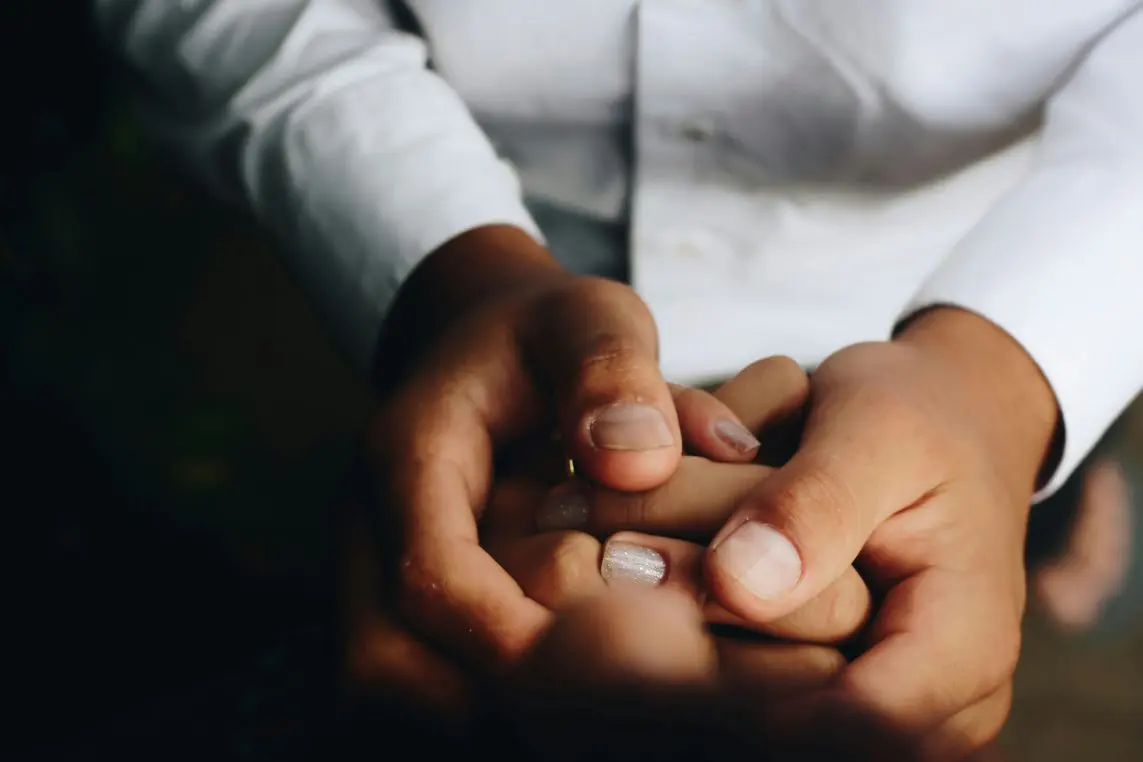 Two adult hands gently clasping another person's fingers, thumb resting on a ringed finger; close-up against a white shirt and dark, blurred background with warm, soft lighting.