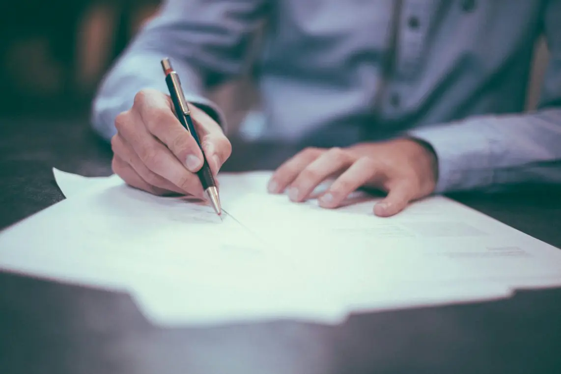 Hand holds black pen, signing or writing on stacked white documents while the other steadies papers on a dark desk; person wears light-blue shirt.