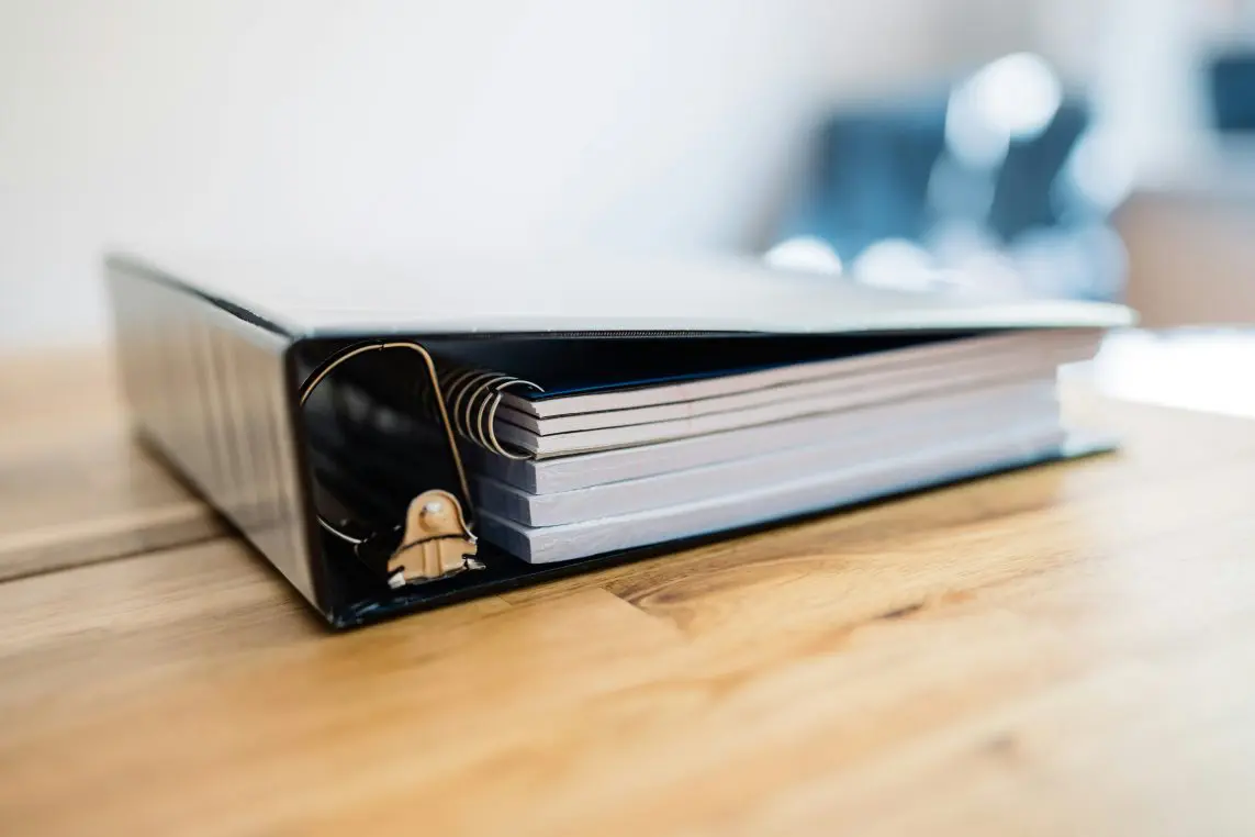 Black three-ring binder rests on a wooden table, its metal rings holding stacked white notebooks; soft daylight illuminates the scene with a blurred office background.
