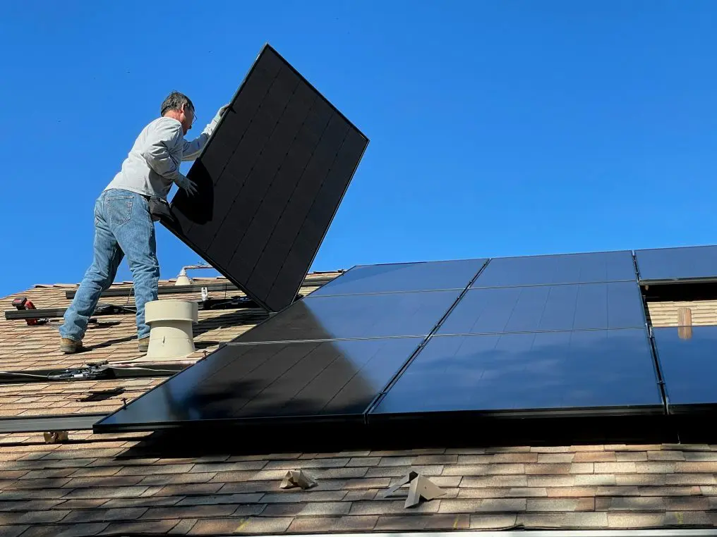 Solar panel being lifted by a worker on a sloped shingled roof, adjacent panels already mounted under a clear blue sky.