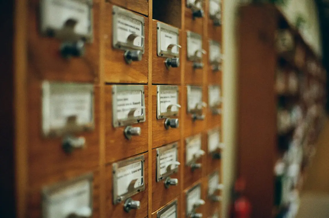 Wooden library card-catalog drawers, each with metal label holders and knobs, arranged in rows and receding into a softly blurred library aisle.