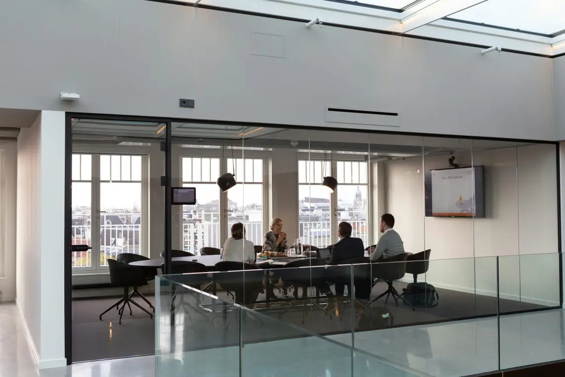 Conference table: five colleagues discuss documents while seated around it in a glass-walled meeting room with pendant lights and large windows showing a city rooftop view.