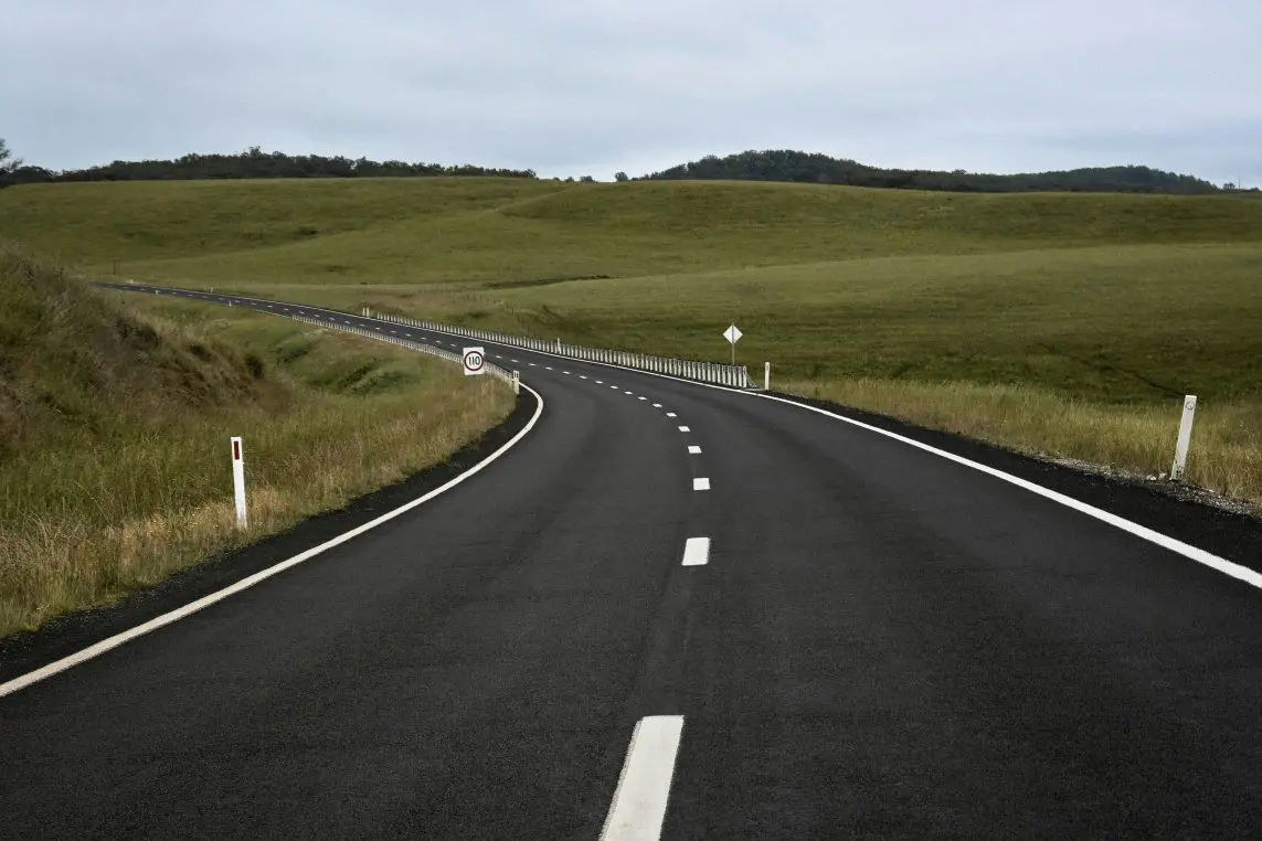 Road curves gently left toward distant hills through open grassy countryside under an overcast sky; white dashed centerline and roadside posts visible. Sign reads 
