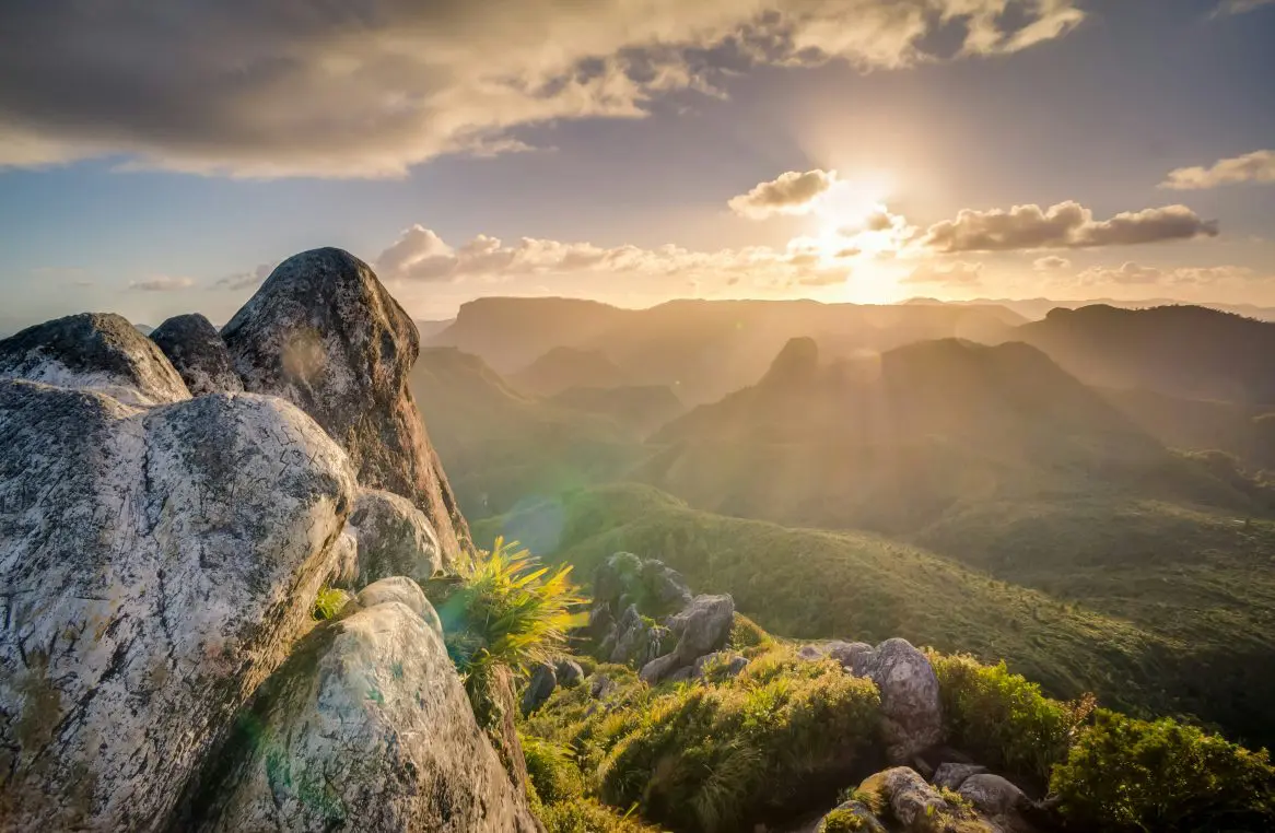 Large weathered rock outcrop catches warm sunlight, overlooking misty, layered mountains as the low sun breaks through clouds, casting golden rays across green valleys.