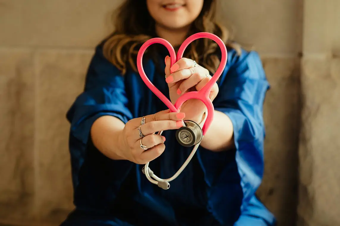 Pink stethoscope shaped into a heart is held toward the camera by a smiling woman wearing a blue dress, seated against a neutral beige couch background.