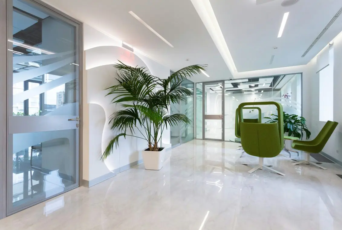 Green swivel lounge chairs sit around a low table in a bright modern office lobby, beside a large potted palm and glass-walled meeting rooms on a glossy marble floor.