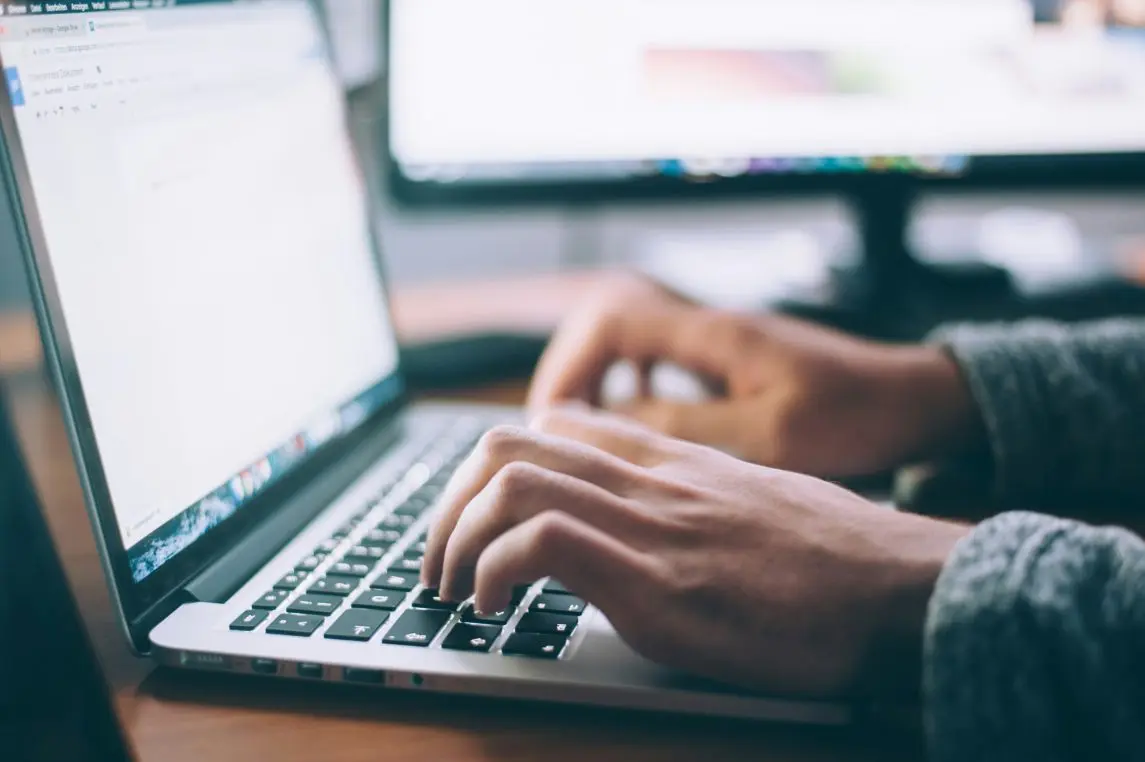 Hands typing on a laptop keyboard, fingers pressing keys while a blurred external monitor glows behind them on a wooden desk, suggesting focused work in a softly lit workspace.