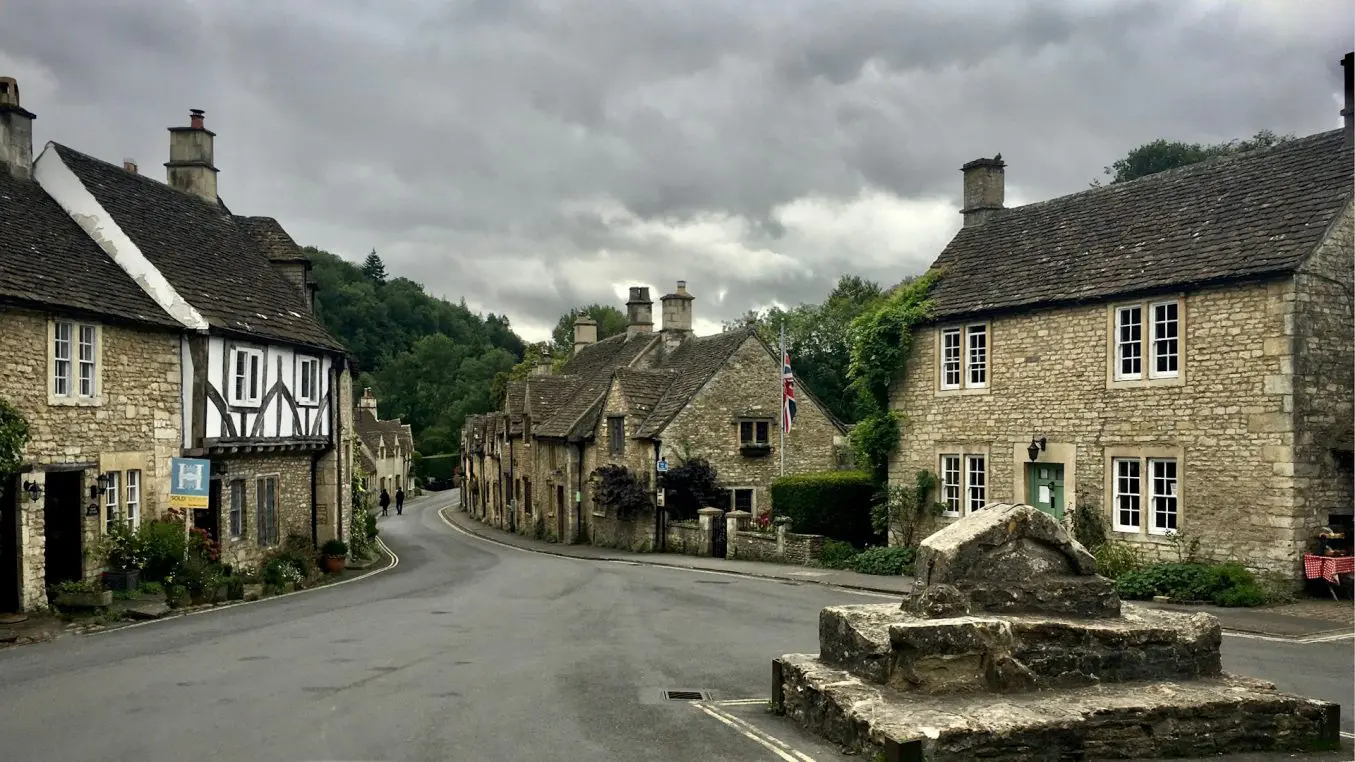 Stone cottages line a gently curving village road, standing quiet under heavy gray clouds, with a low stone monument in the foreground and wooded hills beyond. Visible text: 