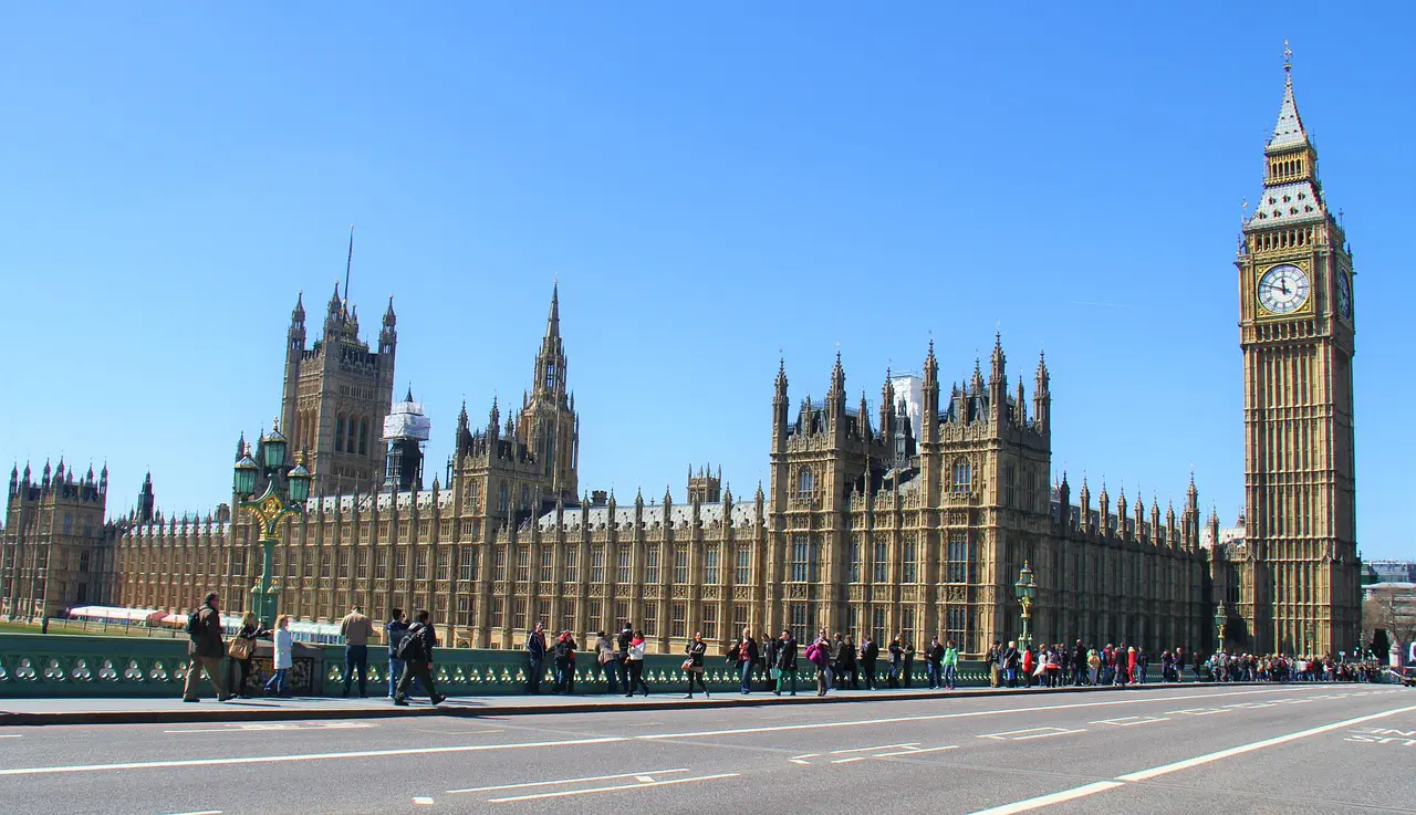 Big Ben and the Palace of Westminster rise beside the River Thames, their Gothic façade bathed in sunlight as pedestrians stroll and gather on Westminster Bridge under clear blue sky.