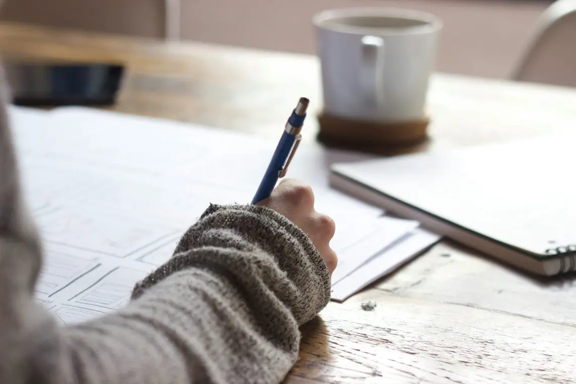 Hand holding a blue pen writes on papers spread across a wooden table, with a spiral notebook and a coffee mug blurred in the background.