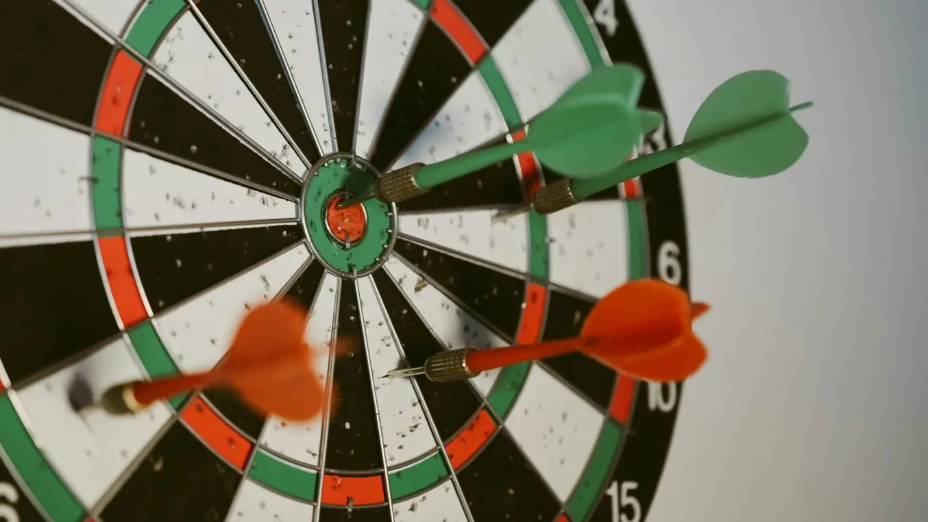 Dartboard — three darts (two green, one orange) embedded near the bullseye; close-up, angled indoor view of a wall-mounted board. Text visible: 
